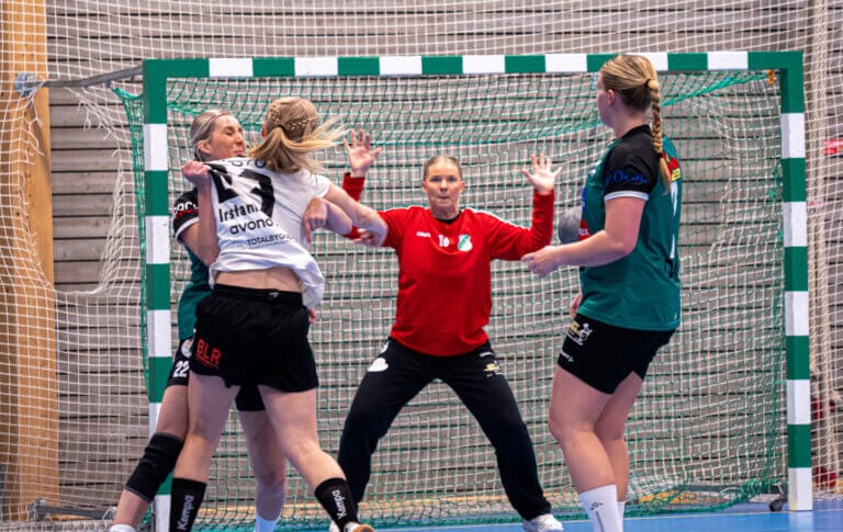 Fyra handbollsspelare från Rimbos damer är i aktion nära målet, och en av dem, iklädd en röd målvaktströja, förbereder sig för att blockera ett skott. Jublet och spänningen är tydlig, med målnätet och planmarkeringarna i bakgrunden.