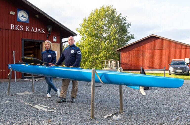 Två personer står bredvid en blå kajak som visas på träställningar framför en röd träbyggnad med texten "RKS Kajak". En automatisk utkast-skylt syns, tillsammans med en annan röd byggnad och en bil på grusytan.