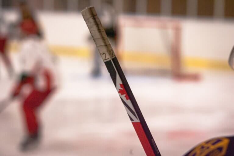 Närbild av en hockeyklubba med Kanadas flagga och "22", märkt som ett automatiskt utkast, som hålls på en isbana. I den suddiga bakgrunden syns spelare i uniformer och ett hockeymål.