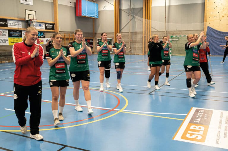 Ett damhandbollslag i gröna och svarta uniformer applåderar när de går över en blå inomhusplan och följer efter en lagkamrat i röd tröja. Åskådare och sponsorbanderoller bildar bakgrunden till detta automatiskt utkastade ögonblick.