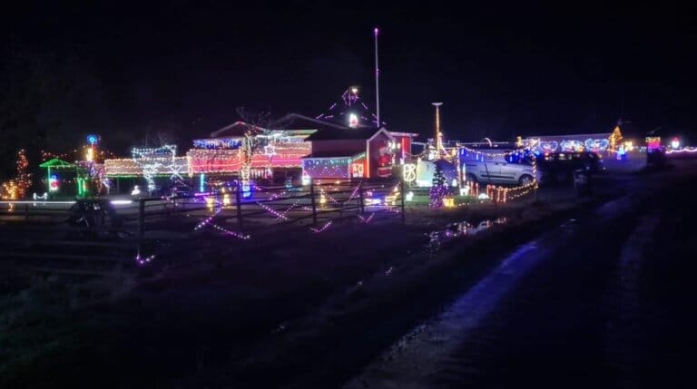 Ett hus och den omgivande gården är dekorerade med färgglada julbelysningar på natten. En pickup är parkerad i närheten och lampor lyser upp staket och byggnader, vilket skapar en festlig atmosfär längs en landsväg.