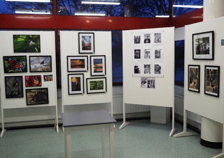 En utställning med inramade fotografier av Hallstaviks fotoklubb är monterad på tre vita vikpaneler i en inomhusutställning. Fotografierna visar landskap, porträtt, abstrakt konst och svartvita bilder. Ett litet grått bord står i mitten.