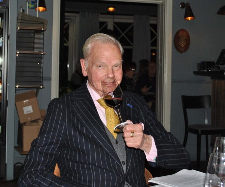 En äldre man i kritstrecksrandig kostym sitter vid ett bord inomhus med ett glas rödvin i handen och ler. I bakgrunden syns kartonger och dekorationer med svenska flaggan.