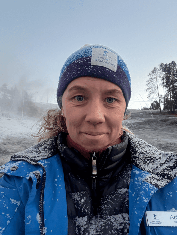 En person i blå jacka, stickad mössa och namnskylt står utomhus en snöig dag. Det är frost på deras kläder och träden i bakgrunden. Himlen är klar och ljus.