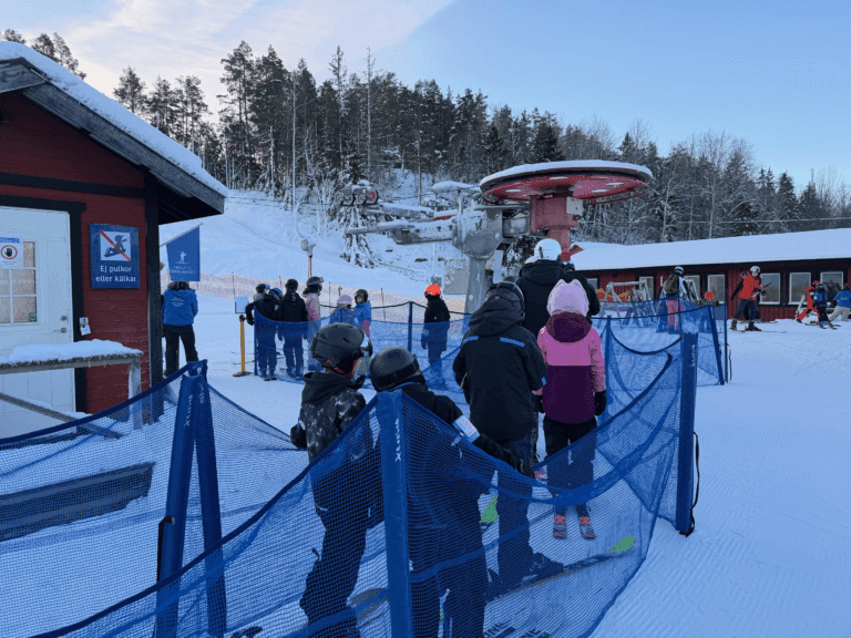 Skidåkare med hjälm och vinterutrustning står i kö bakom ett blått nät vid en skidliftstation en snöig dag, med byggnader och träd i bakgrunden.