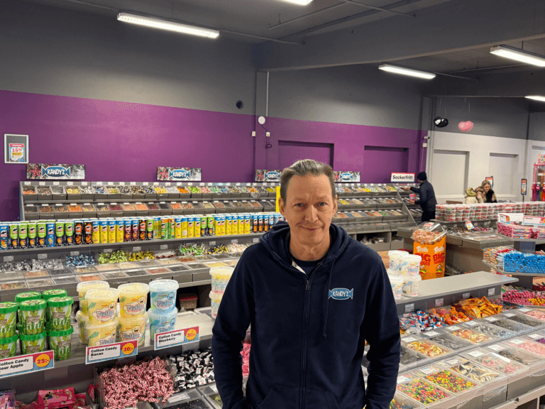 En man i marinblå huvtröja står inne i en starkt upplyst godisbutik, med hyllor och backar fyllda med olika färgglada godisbitar, drycker och snacks bakom sig. Väggarna är målade i lila och grått.