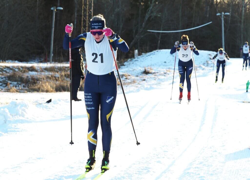 Saga Westerlund, med nummerlapp märkt 21, tävlar i ett längdskidlopp under tävlingar Falun i ett snöigt spår, följd av andra skidåkare. I utomhusmiljön finns träd och partier med barmark.