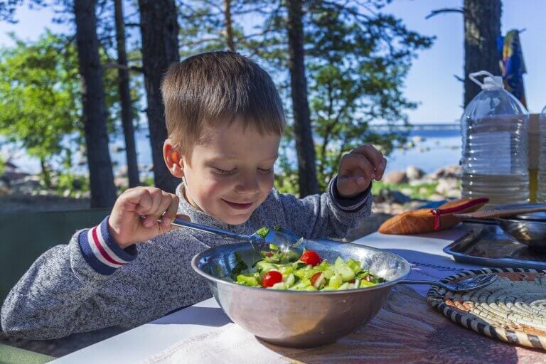 En ung pojke sitter vid ett bord utomhus och ler medan han använder sked och gaffel för att äta en sallad med körsbärstomater och gurkor. Träd, en sjö och picknickutrustning omger honom i bakgrunden.