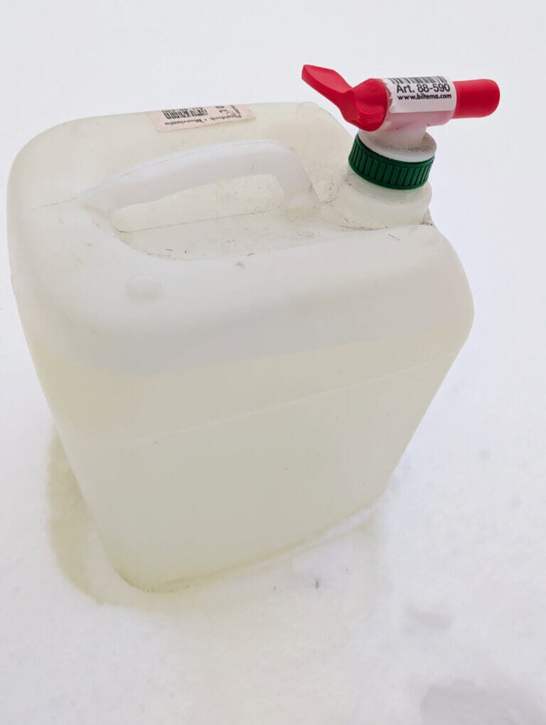 En vit plastbehållare med röd och vit pip står på en snöfläck. Behållaren har ett grönt skruvlock och en etikett med text på toppen. Behållaren ser ut att innehålla vätska.