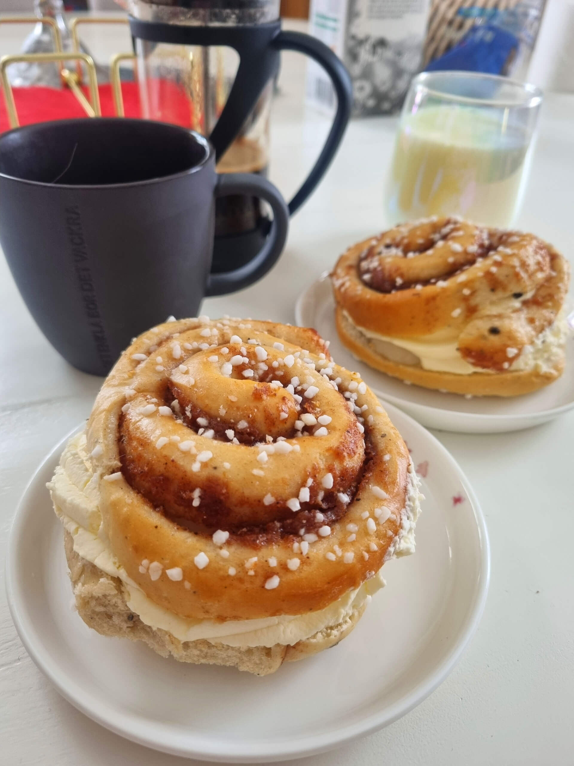 Två kanelbullar fyllda med grädde serveras på små vita tallrikar. En svart mugg, ett glas mjölk, en kaffepanna och olika köksredskap syns i bakgrunden på ett vitt bord.