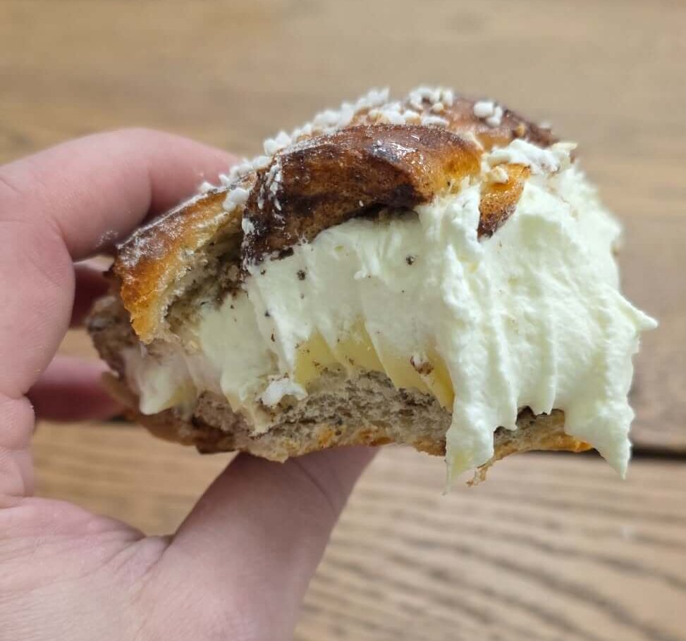 En hand som håller i en bakelse fylld med vispgrädde och strösocker på toppen, över ett träbord.