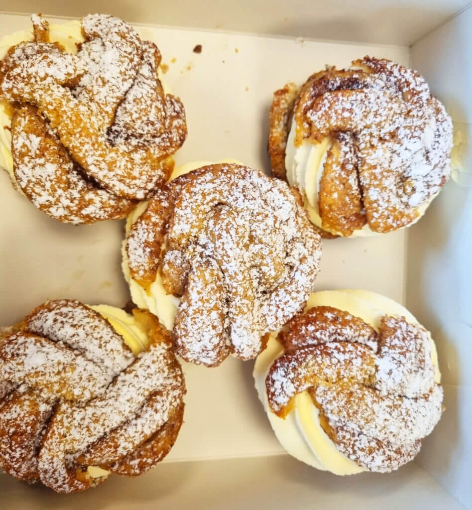 Fem gräddfyllda bakverk toppade med strösocker är arrangerade i en vit låda. Bakverken har ett vridet, gyllenbrunt utseende med synliga lager och virvlar av gräddfyllning.