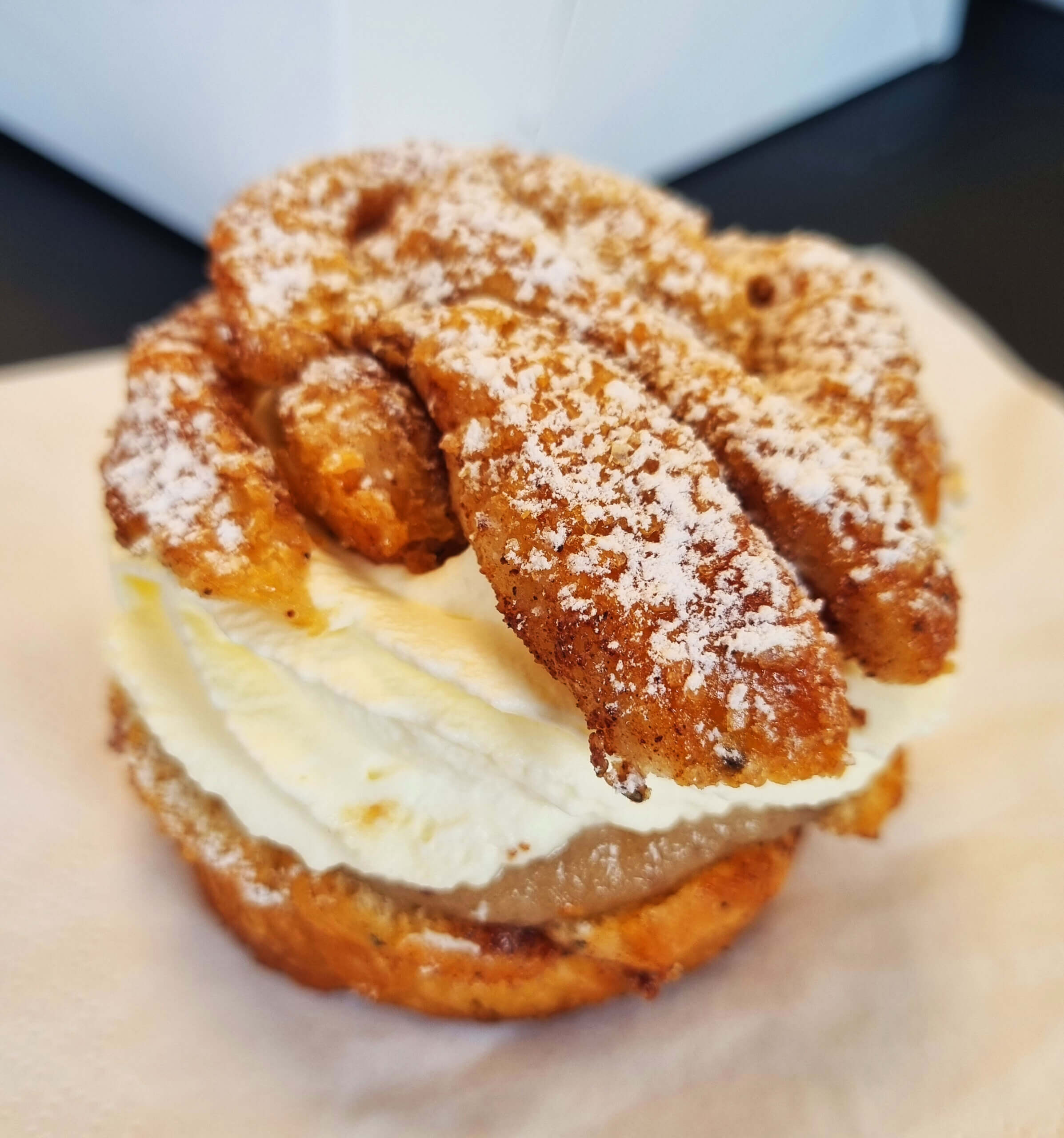 En gräddfylld bakelse toppad med strösocker ligger på en vit servett. Bakelsen har ett gyllenbrunt, strukturerat yttre och är fylld med spritsade lager av grädde och en ljusbrun fyllning.