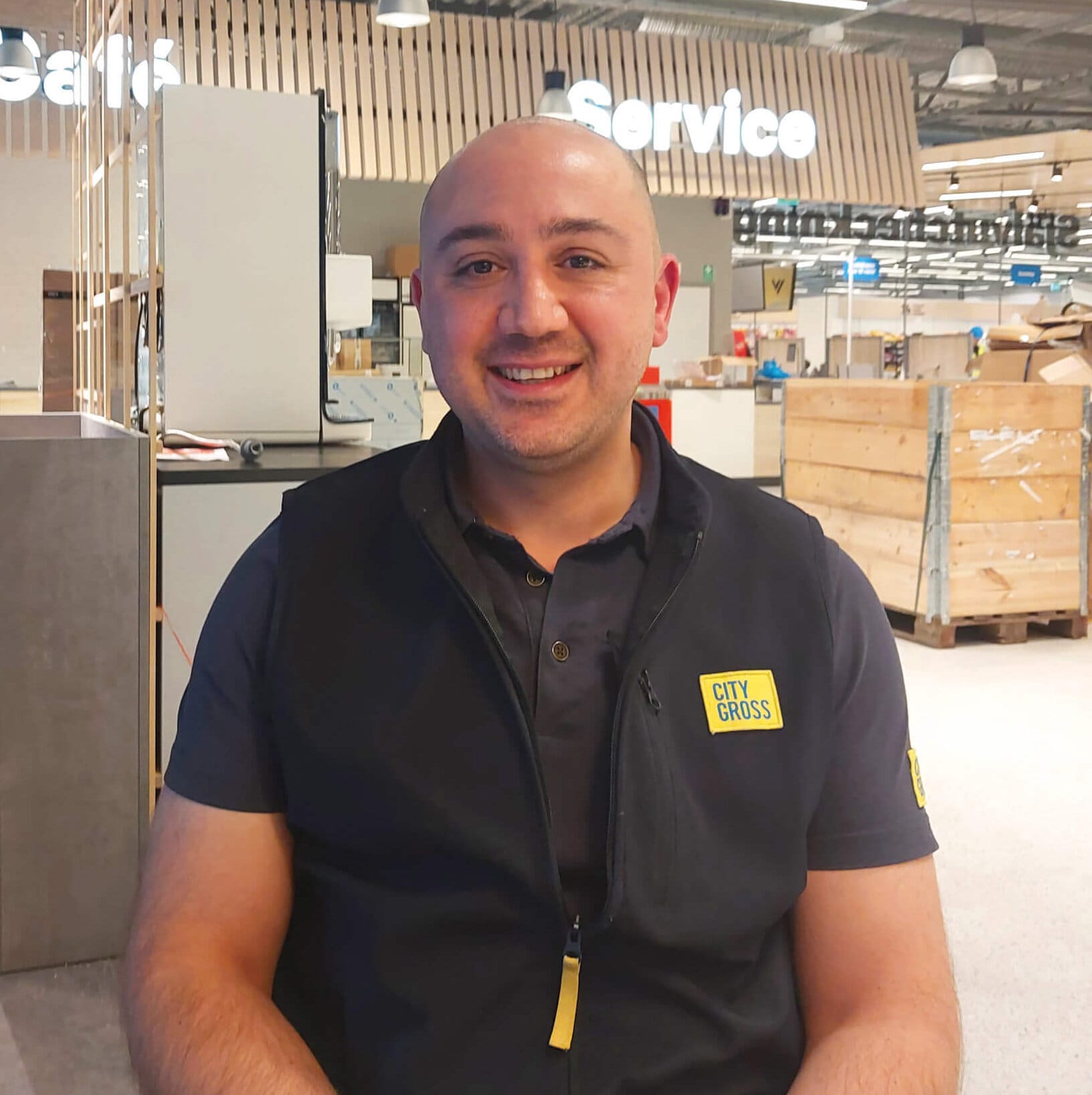 En man sitter och ler i en butik med synliga skyltar med texten "Café" och "Service" i bakgrunden. Han bär en mörk väst och en gul bricka där det står "City Gross". Butiksinredningen är ljus och modern.