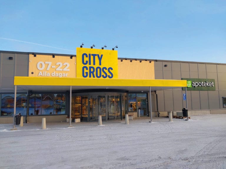 Ingången till en City Gross stormarknad med en gul skylt, bredvid ett Apoteket-apotek. Snö täcker marken och parkeringsplatsen. Öppettiderna "07-22 Alla dagar" visas ovanför entrén.