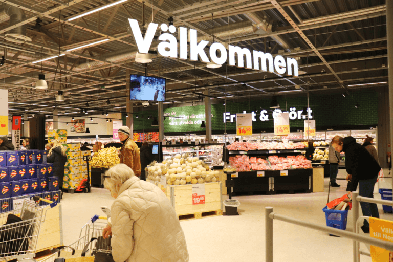 Interiör från en stormarknad med kunder som skjuter på vagnar, varuställ och skyltar på svenska, inklusive en stor "Välkommen"-skylt som hänger från taket. Stark belysning och staplade varor syns.