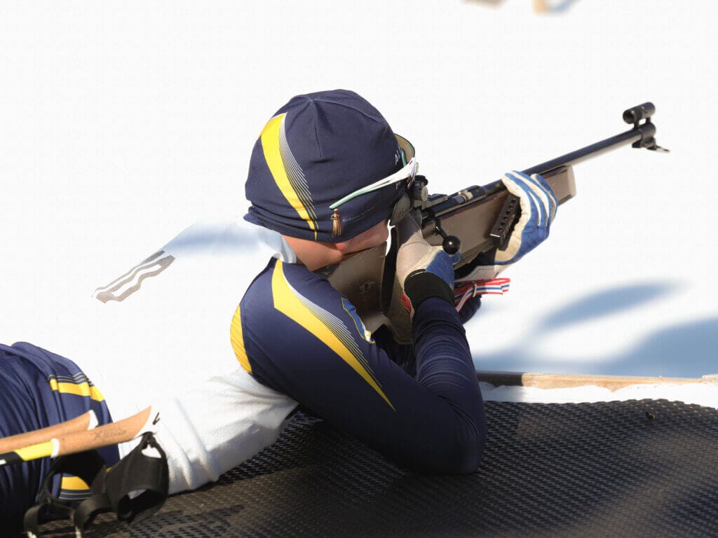 En person i blågul dräkt ligger på mage på en matta och siktar med ett gevär mot ett mål. Den snöiga miljön tyder på ett imponerande framträdande vid ett skidskytteevenemang, möjligen av Häverödals skidskyttar.