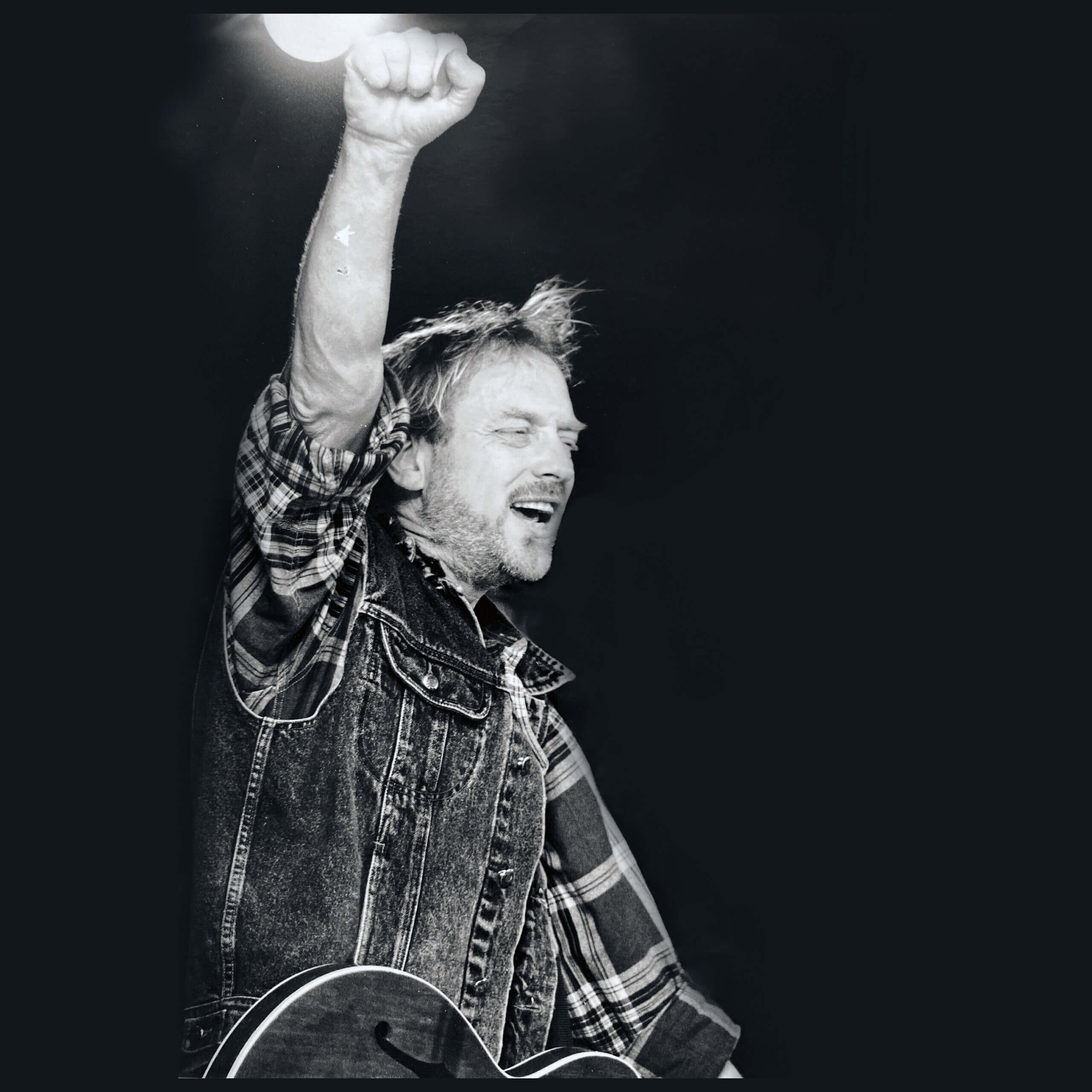 En man i rutig skjorta och jeansväst håller upp högerarmen med en knuten näve medan han spelar på en akustisk gitarr. Han har ett fokuserat uttryck och är upplyst mot en mörk bakgrund.
