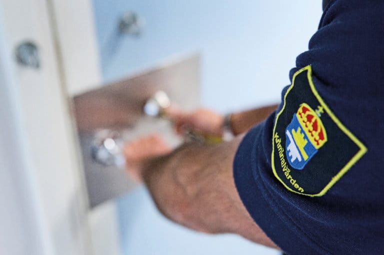 En person i uniform med Kriminalvården-märke låser upp eller låser en metallucka i en dörr, möjligen på en kriminalvårdsanstalt eller ett häkte. Fokus ligger på uniformen och armen.