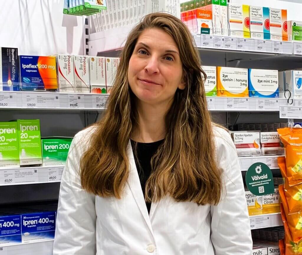 En kvinna med långt brunt hår och vit labbrock står framför hyllor med olika mediciner och hälsoprodukter på ett apotek eller en droghandel.