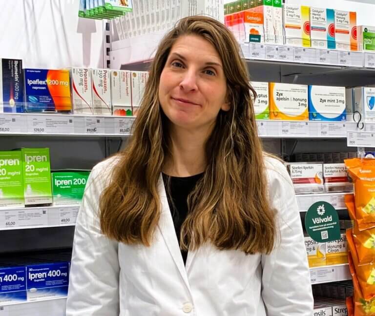 En kvinna med långt brunt hår och vit labbrock står framför hyllor med olika mediciner och hälsoprodukter på ett apotek eller en droghandel.