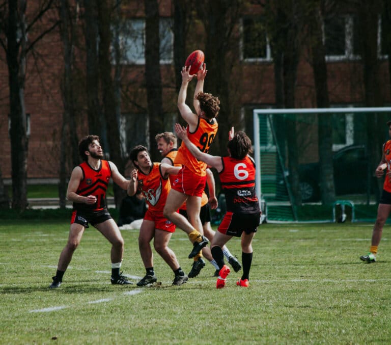 Fyra idrottare i orange och svarta uniformer tävlar om att få bollen under en utomhusmatch på en gräsplan. En spelare hoppar högt med armarna i luften, medan de andra tittar på eller förbereder sig för att fånga bollen. Träd och en byggnad syns i bakgrunden.