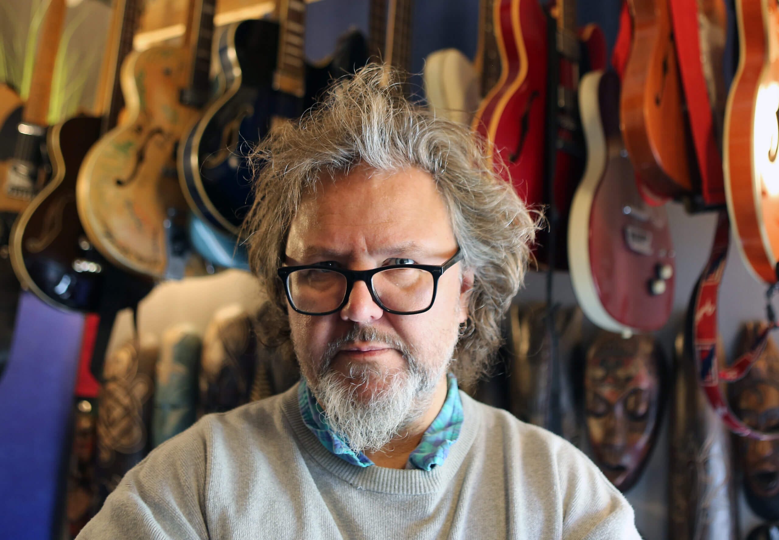 En medelålders man med glasögon och grått hår står inomhus, klädd i en ljus tröja och rutig skjorta. På väggen bakom honom hänger flera elektriska och akustiska gitarrer.