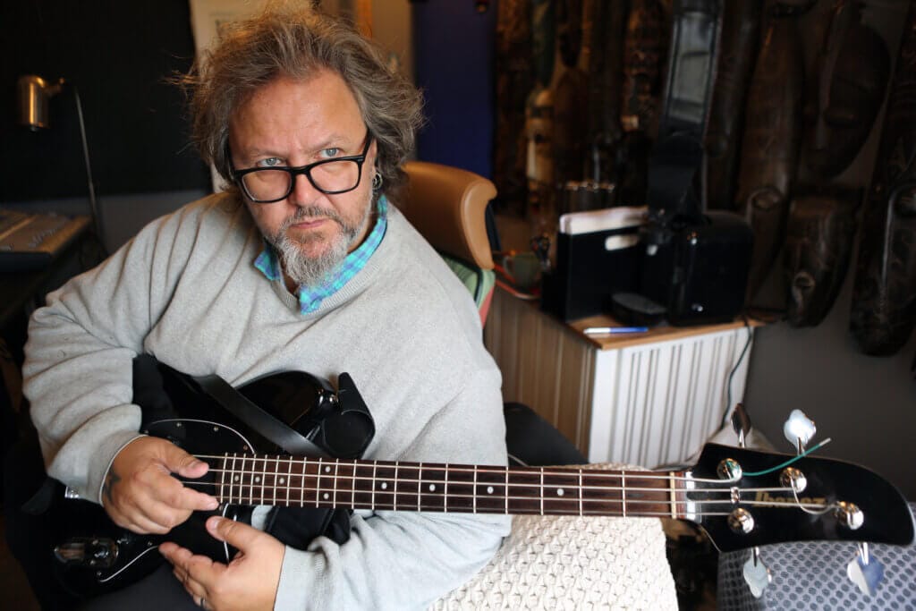 En person med grått hår och glasögon sitter inomhus och håller i en svart elbasgitarr. Personen tittar in i kameran och är klädd i en ljusgrå tröja över en blårutig skjorta. I bakgrunden syns musik- och kontorsutrustning.