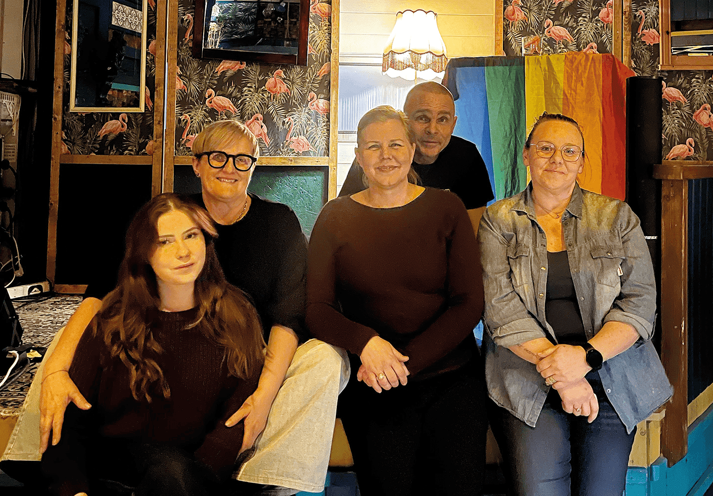 Fem vuxna poserar för ett gruppfoto inomhus framför en färgglad vägg med speglar och ett flamingomönster. Fyra sitter, en står bakom dem. I bakgrunden syns en lampa och en regnbågsflagga.