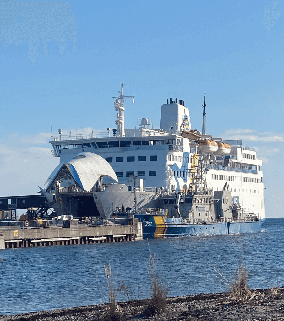En stor färja ligger vid en pir bredvid ett mindre blågult fartyg. Fartygets ramp är nedfälld på kajen och livbåtarna syns på de övre däcken under en klarblå himmel.