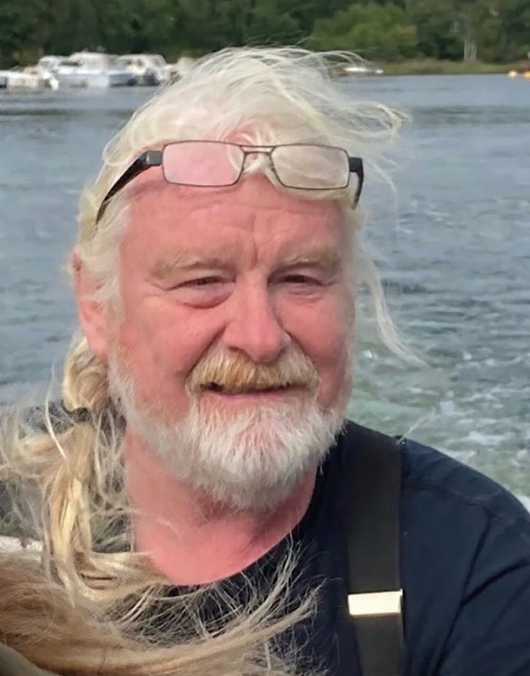 En äldre man med långt vitt hår och skägg, med glasögon på huvudet och mörk skjorta, sitter på en båt med vatten och strandlinje i bakgrunden.