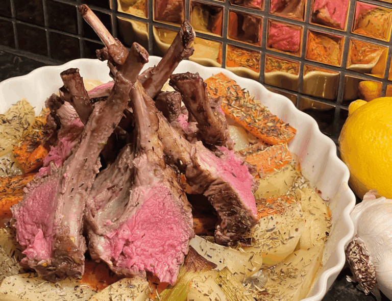 En lammstek som tillagats medium rare är placerad ovanpå skivade rostade grönsaker i en vit skål, med citron, vitlök och en speglad kakelplatta synlig i bakgrunden.