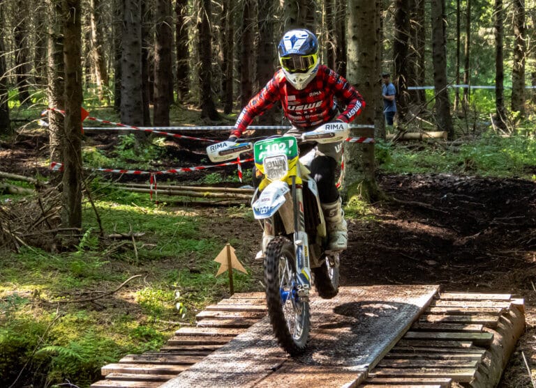 En person iförd skyddsutrustning och en röd och svart tröja kör en crossmotorcykel över en smal träbro i en skog under ett terränglopp med sikte på dubbla pallplatser som Wiggo Lifendahl i Spanien.