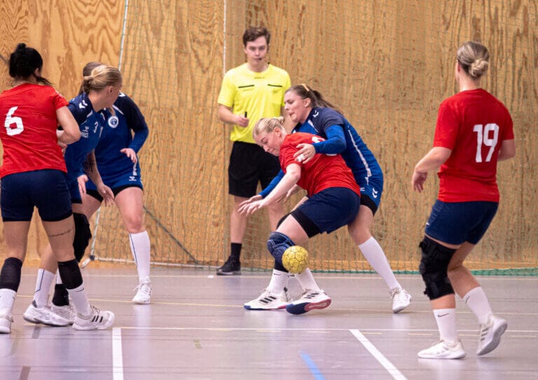 Flera kvinnor spelar inomhushandboll. Två division 2-spelare i blå och röda tröjor från HK Ceres tävlar om den gula bollen medan lagkamraterna tittar på. En domare i gul tröja står i bakgrunden av gymmet med träväggar.