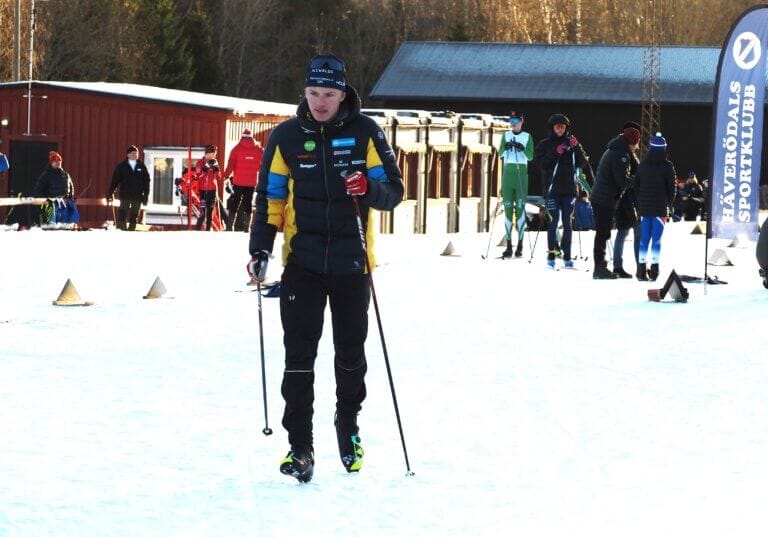 En person i vintersportkläder står på snö med skidstavar, omgiven av andra och byggnader på Häverödals. En banderoll och träd syns i bakgrunden, vilket tyder på att scenen är från ett skidevenemang som JSM Guld eller en tävling.