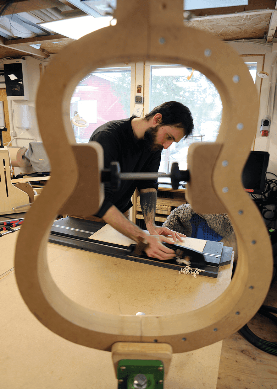 En man arbetar på en träbit vid en arbetsbänk i en verkstad, sedd genom en stor gitarrformad ram i förgrunden. Verktyg och träbearbetningsmaterial syns runt omkring honom.