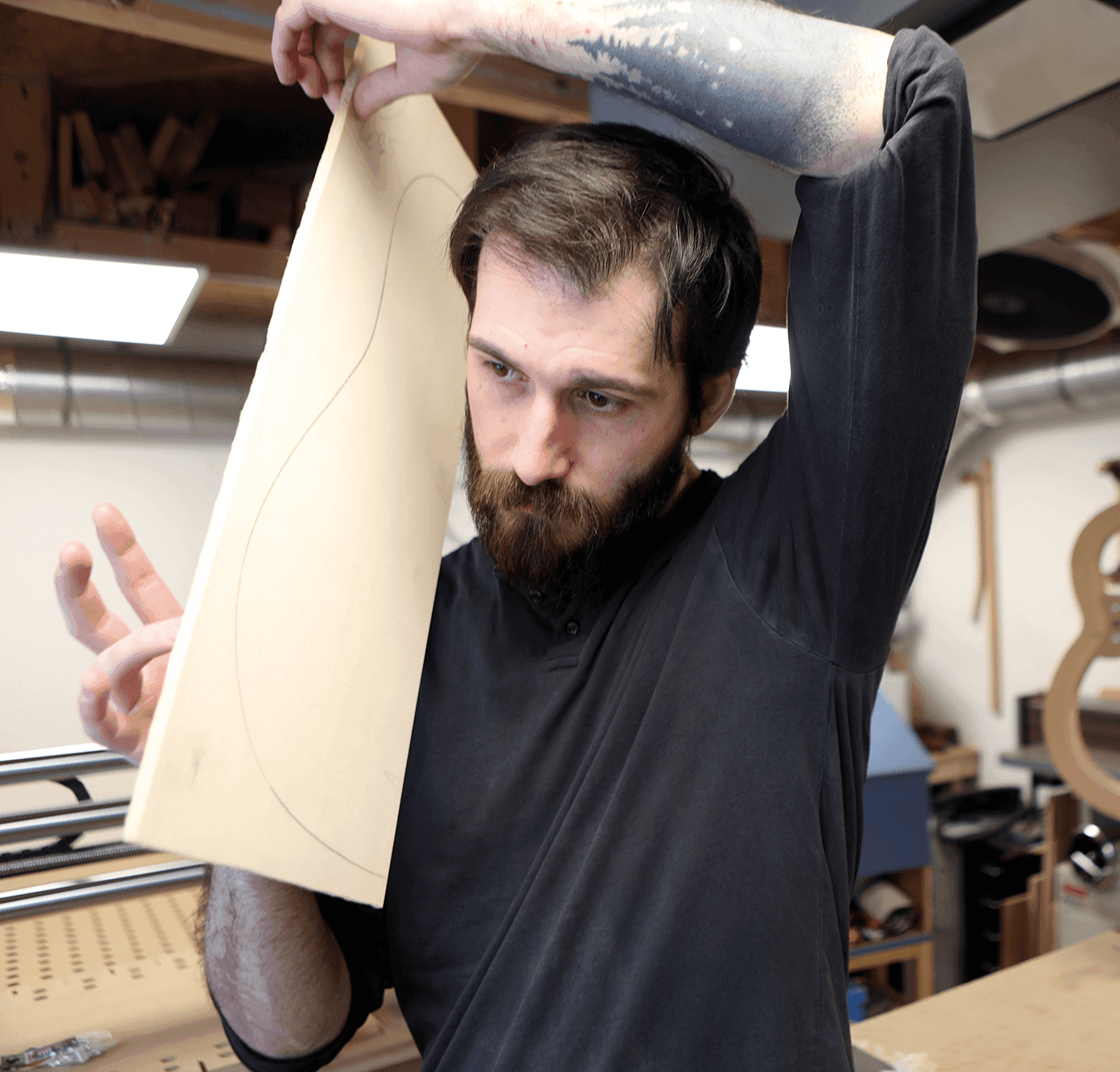 En man i en verkstad håller en träskiva med en böjd linje på och lyfter den under armen. Han har skägg, bär en mörk skjorta och står nära träbearbetningsverktyg och material.
