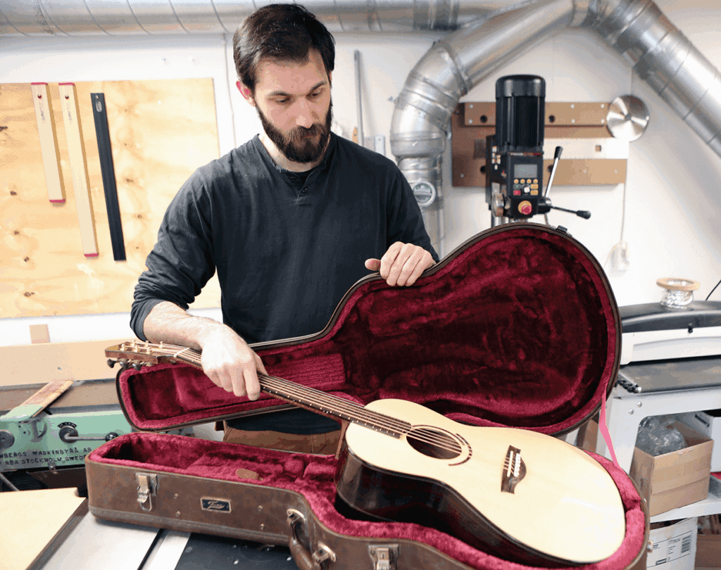 En man med skägg placerar en ljus akustisk gitarr i ett gitarrfodral med rött sammetsklädsel i en verkstad med olika verktyg och utrustning i bakgrunden.