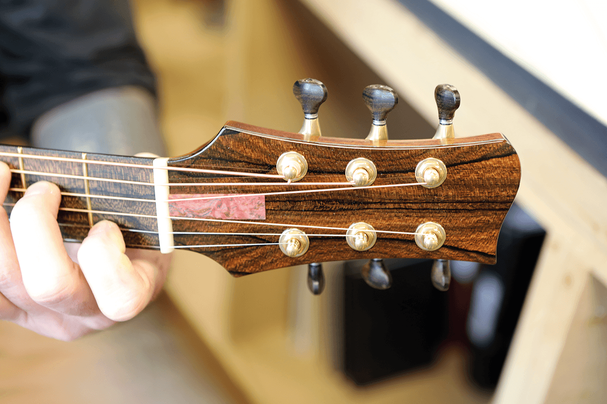 Närbild av en persons vänstra hand som håller i halsen på en akustisk gitarr, med huvuddocka, stämskruvar och strängar i trä, i en väl upplyst inomhusmiljö.