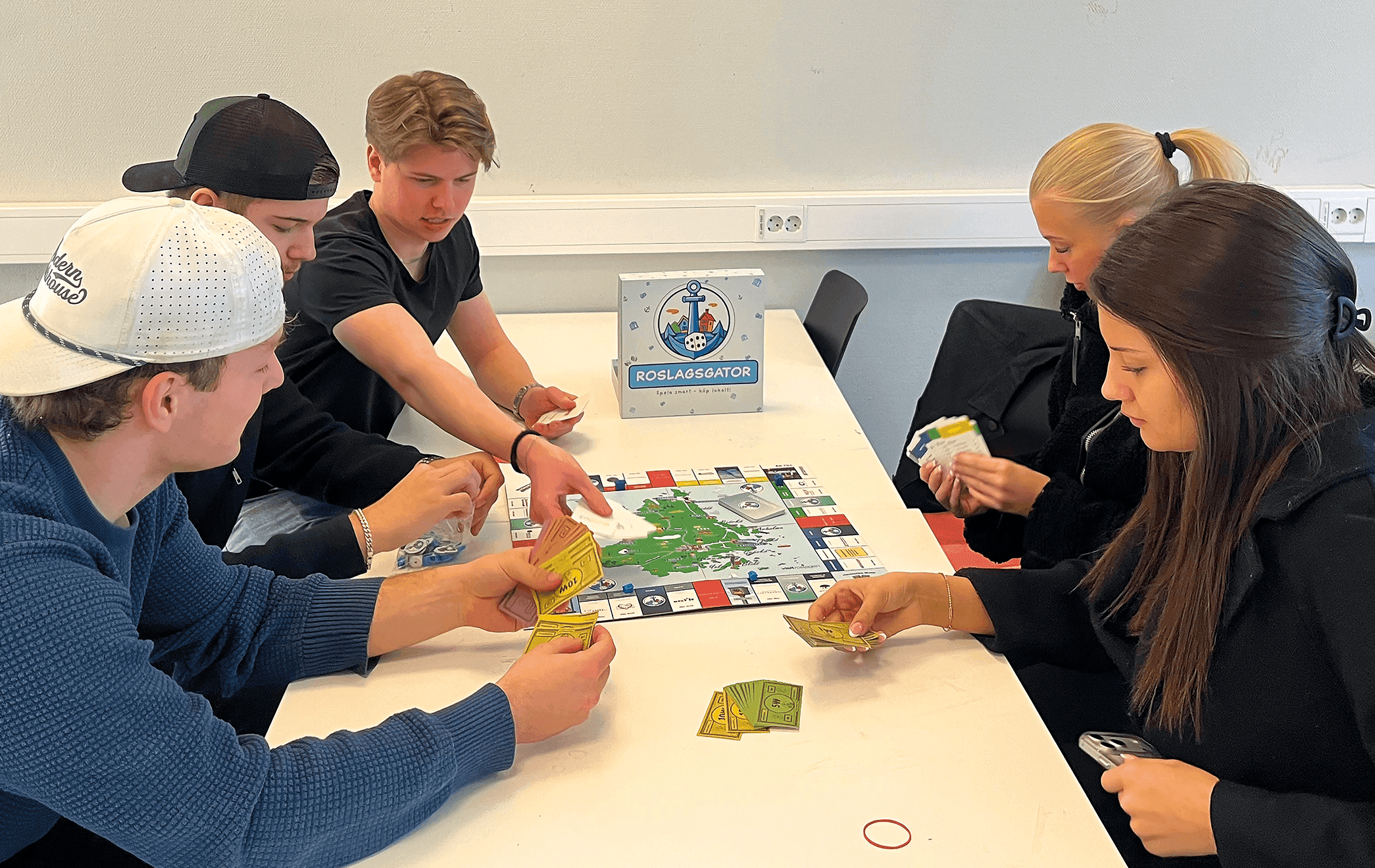 Fem personer sitter runt ett bord och spelar ett brädspel. Fyra av dem håller i kort och en flyttar en pjäs på spelbrädet. Bordet är vitt och väggen bakom dem är ljusgrå.