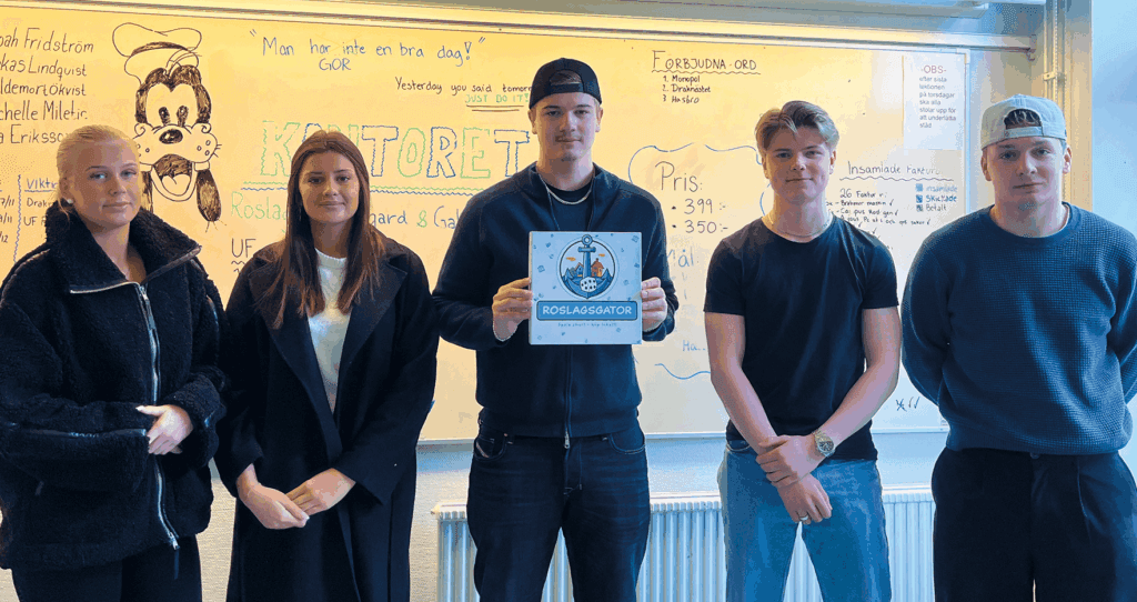 Fem unga vuxna står inomhus framför en whiteboardtavla med handskriven text och teckningar. Personen i mitten håller i en skylt med en blå kant och en logotyp på. Alla tittar in i kameran.