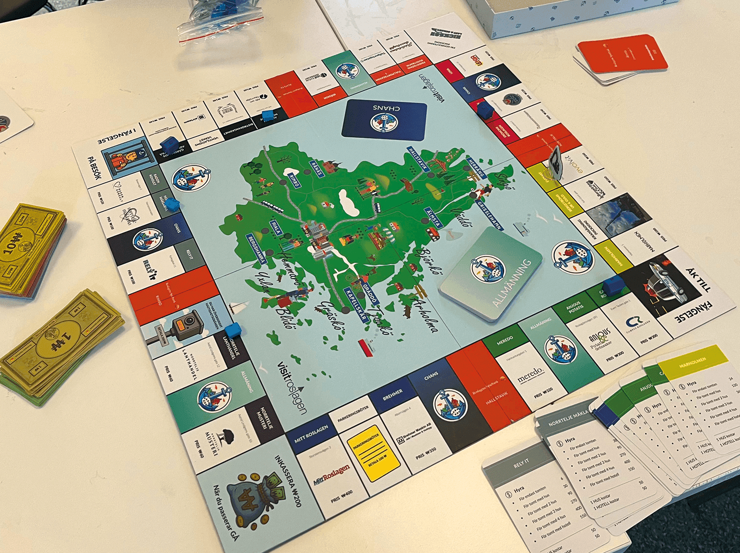 Ett brädspel står uppställt på ett vitt bord med färgglada kort, papperspengar och spelpjäser. Spelbrädet har en karta i mitten och olika märkta utrymmen runt kanterna. Spellådan och komponenterna är synliga.