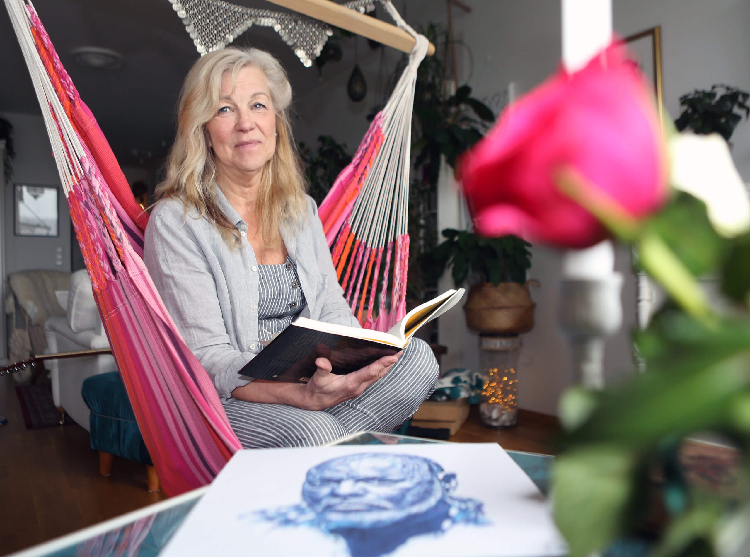 En kvinna med långt grått hår sitter i en rosa hängmatta inomhus och håller i en uppslagen bok. En suddig rosa ros och en skiss av ett ansikte syns i förgrunden. Rummet innehåller växter och mjuk belysning.