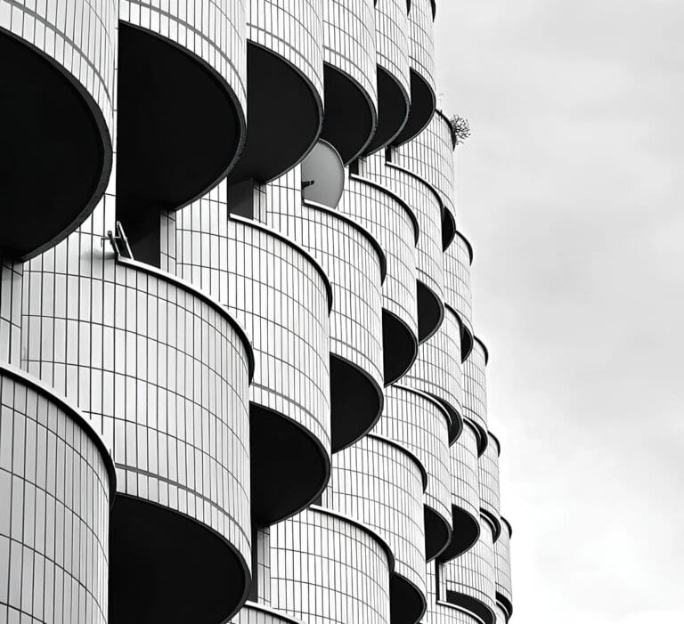 Närbild av en modern byggnadsfasad med upprepade svängda balkonger täckta av vitt kakel, arrangerade i ett förskjutet vertikalt mönster mot en molnig himmel.