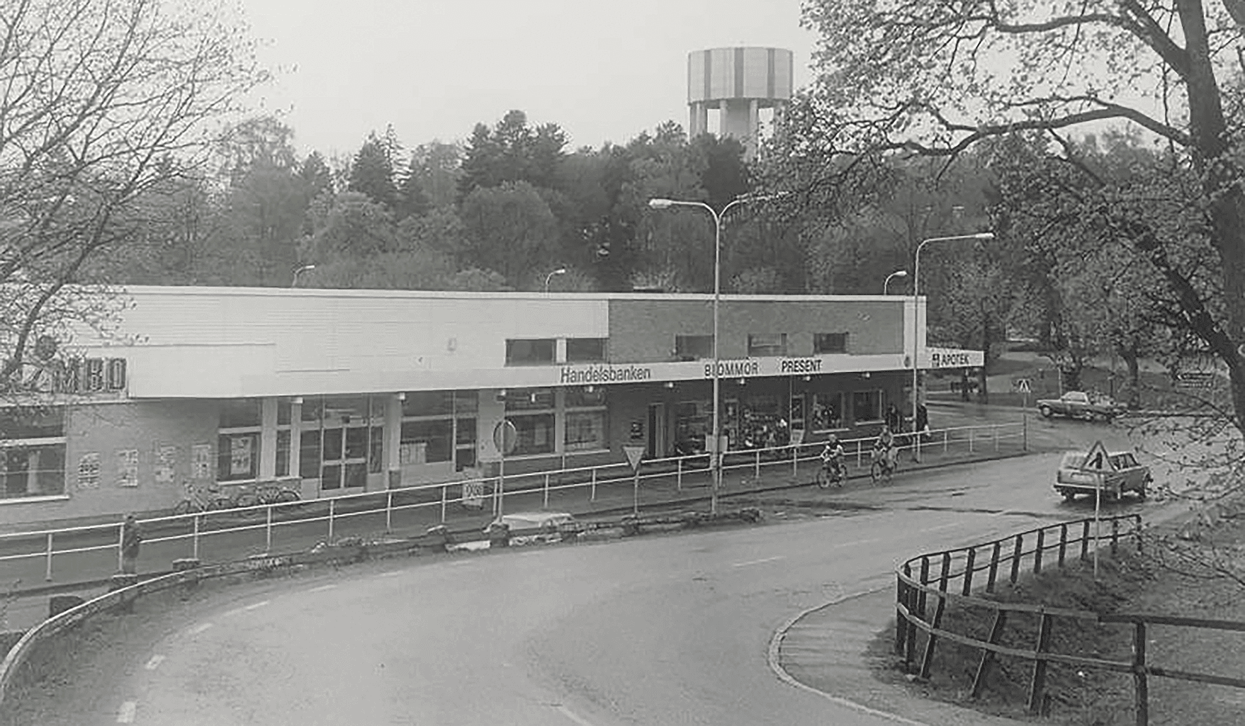 Ett svartvitt foto visar ett litet köpcentrum med skyltfönster, en krökt väg framför, en bil parkerad i närheten, cyklar och träd i bakgrunden. Ett vattentorn syns ovanför trädgränsen.