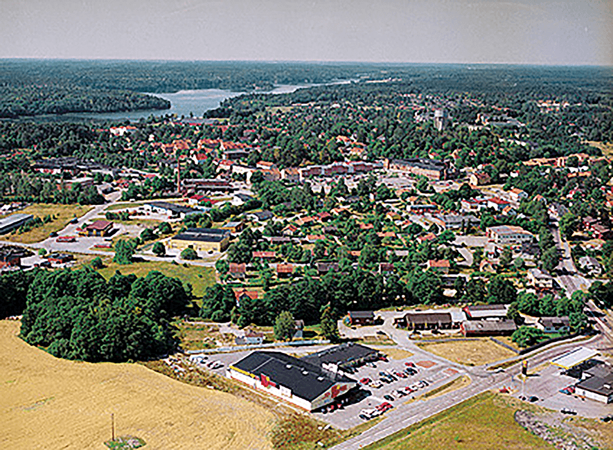 Flygfoto över en liten stad omgiven av tät skog och en sjö i fjärran, med bostadshus, vägar och några kommersiella byggnader synliga i förgrunden.