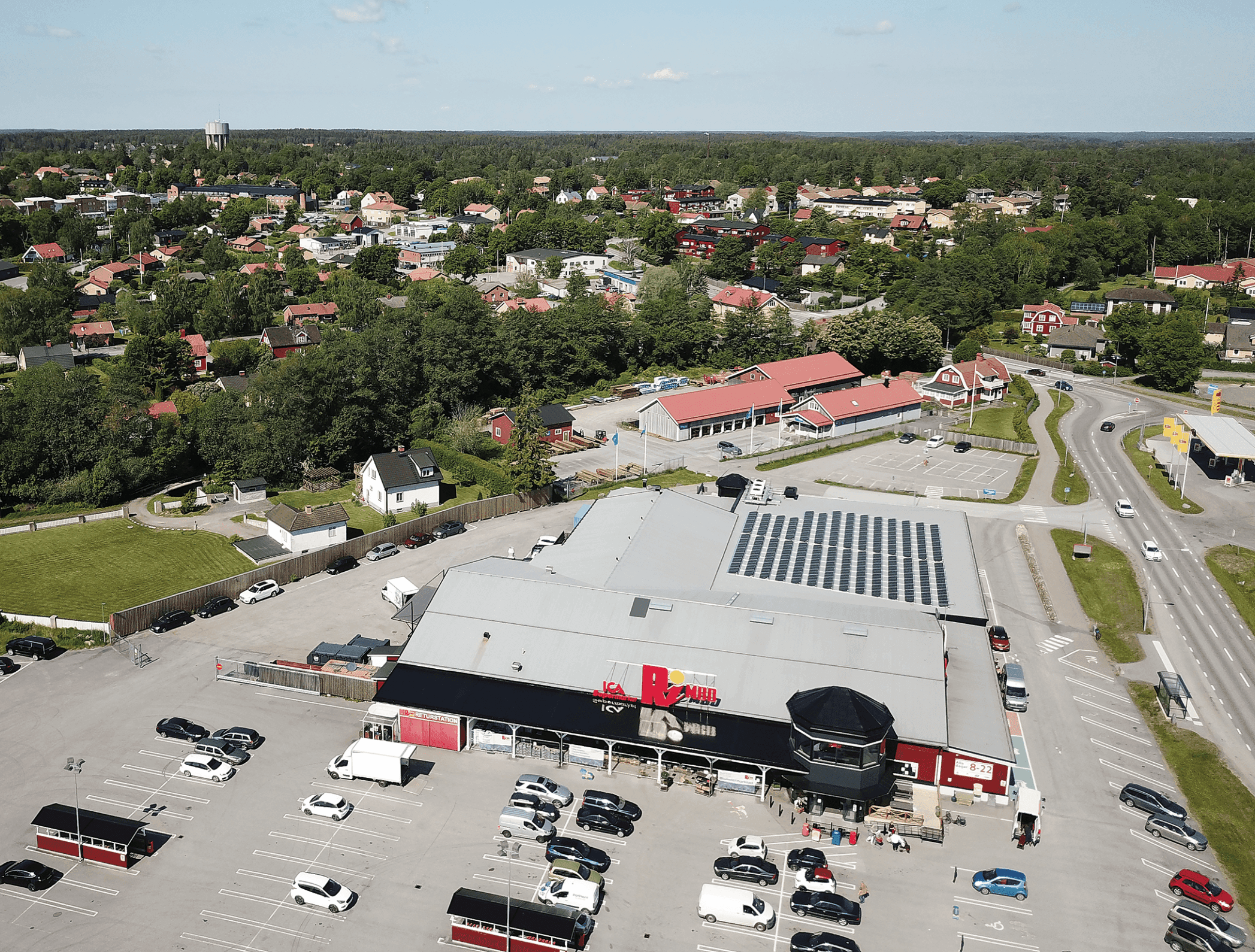 En stor parkeringsplats med många bilar omger en kommersiell byggnad märkt "Berns". I området finns vägar, mindre byggnader, träd och en stadsbild i fjärran under en klarblå himmel.