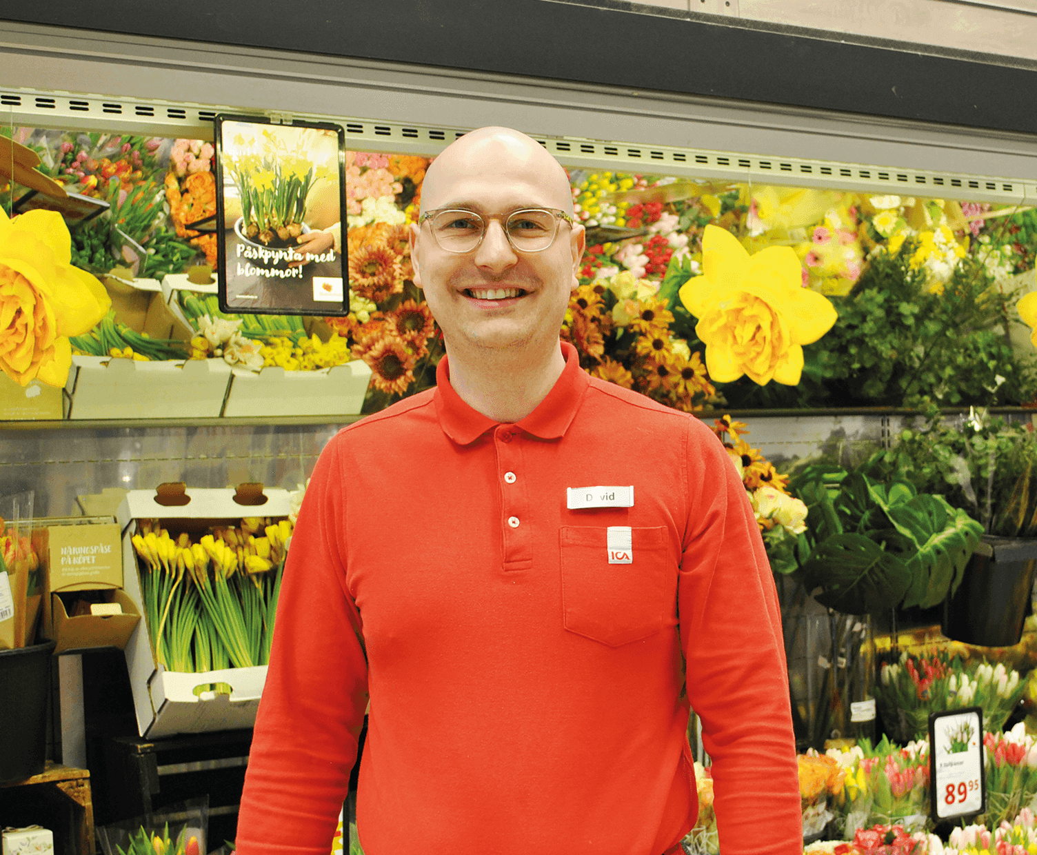 En person i röd skjorta och glasögon står framför en kyldisplay fylld med olika buketter och arrangemang av gula och flerfärgade blommor i en butik.