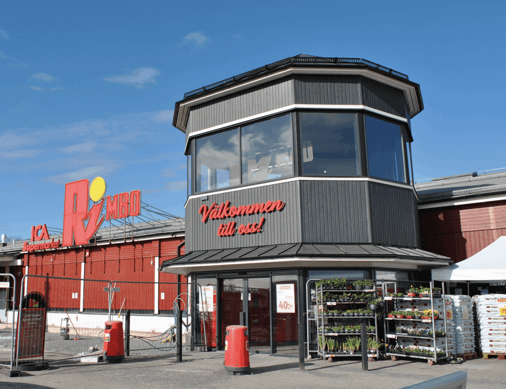 En röd ICA Kvantum Rimbo livsmedelsbutik med en stor skylt, en åttkantig entré, byggmaskiner, stängsel och varuhögar utanför en klar dag.