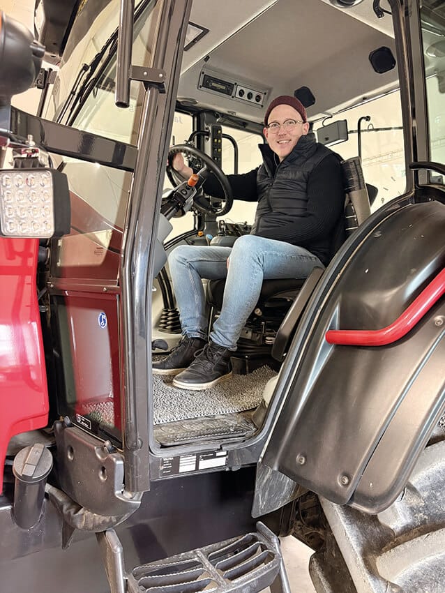 En person med glasögon, rödbrun mössa, svart väst och jeans sitter i förarsätet på en röd och svart traktor, ler och håller i ratten i en väl upplyst inomhusmiljö.
